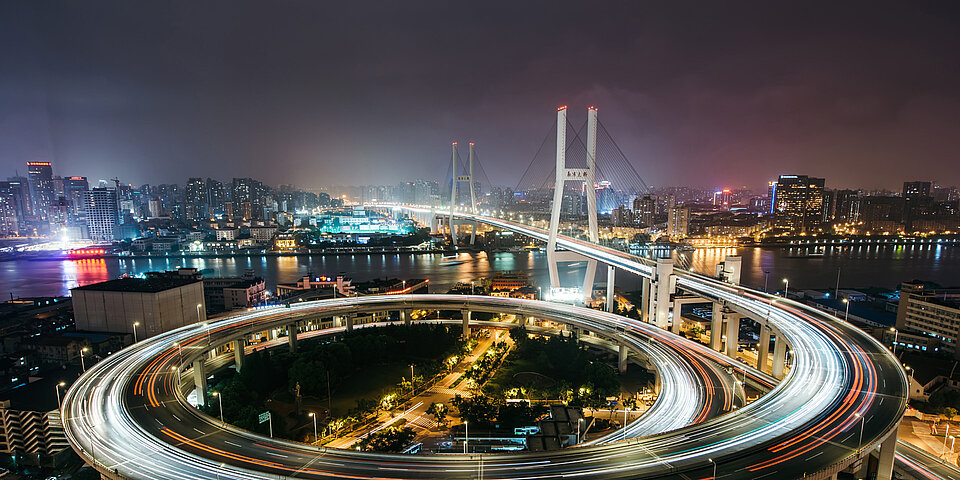 Nachtansicht einer belebten Stadt mit beleuchteter Brücke und kreisförmigem Verkehrskreisel, der von Lichtspuren durchzogen ist.