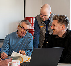 Vier Männer besprechen lächelnd Dokumente vor einem Laptop in einem entspannten Büroumfeld.